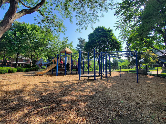A playground at Hereford Park, with blue climbing equipment and a slide beneath leafy trees.